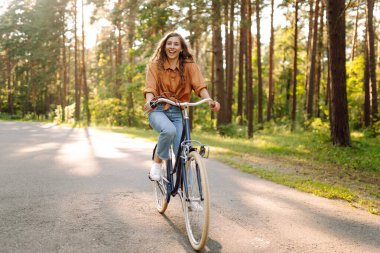 Yoğun güneşli bir ormanda bisiklet süren genç bir kadın. Doğanın tadını çıkaran ve gün batımında bisiklet süren mutlu bir kadın. Hafta sonu konsepti. Etkin yaşam biçimi.