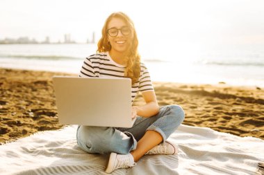 Günbatımı plajında dizüstü bilgisayarı olan genç bir kadın. Günlük kıyafetleriyle deniz manzarasından hoşlanan ve dizüstü bilgisayarla çalışan serbest çalışan bir kadın. Doğa ve teknoloji kavramı. Yaşam biçimi.