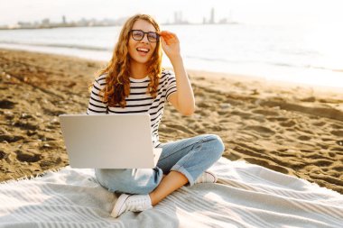 Günbatımı plajında dizüstü bilgisayarı olan genç bir kadın. Günlük kıyafetleriyle deniz manzarasından hoşlanan ve dizüstü bilgisayarla çalışan serbest çalışan bir kadın. Doğa ve teknoloji kavramı. Yaşam biçimi.