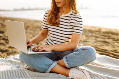 Günbatımı plajında dizüstü bilgisayarı olan genç bir kadın. Günlük kıyafetleriyle deniz manzarasından hoşlanan ve dizüstü bilgisayarla çalışan serbest çalışan bir kadın. Doğa ve teknoloji kavramı. Yaşam biçimi.