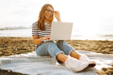 Günbatımı plajında dizüstü bilgisayarı olan genç bir kadın. Günlük kıyafetleriyle deniz manzarasından hoşlanan ve dizüstü bilgisayarla çalışan serbest çalışan bir kadın. Doğa ve teknoloji kavramı. Yaşam biçimi.