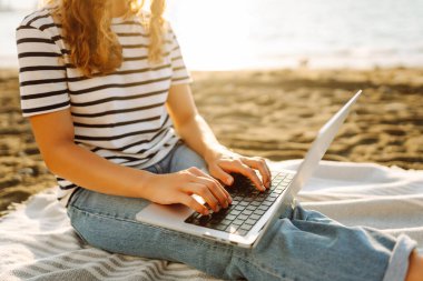 Günbatımı plajında dizüstü bilgisayarı olan genç bir kadın. Günlük kıyafetleriyle deniz manzarasından hoşlanan ve dizüstü bilgisayarla çalışan serbest çalışan bir kadın. Doğa ve teknoloji kavramı. Yaşam biçimi.