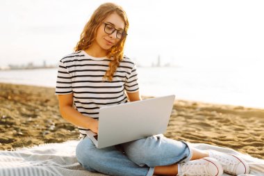 Günbatımı plajında dizüstü bilgisayarı olan genç bir kadın. Günlük kıyafetleriyle deniz manzarasından hoşlanan ve dizüstü bilgisayarla çalışan serbest çalışan bir kadın. Doğa ve teknoloji kavramı. Yaşam biçimi.