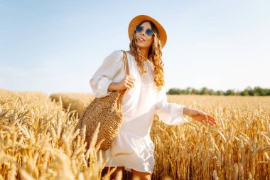 Şapkalı güzel bir kadın gün batımında altın buğday tarlasında yürüyor. Beyaz elbiseli mutlu kadın kırsal bir manzaranın tadını çıkarıyor. Güzellik, moda, hafta sonu konsepti. Yaşam biçimi.