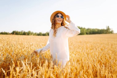 Şapkalı güzel bir kadın gün batımında altın buğday tarlasında yürüyor. Beyaz elbiseli mutlu kadın kırsal bir manzaranın tadını çıkarıyor. Güzellik, moda, hafta sonu konsepti. Yaşam biçimi.