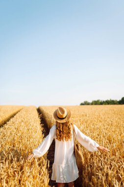 Şapkalı güzel bir kadın gün batımında altın buğday tarlasında yürüyor. Beyaz elbiseli mutlu kadın kırsal bir manzaranın tadını çıkarıyor. Güzellik, moda, hafta sonu konsepti. Yaşam biçimi.