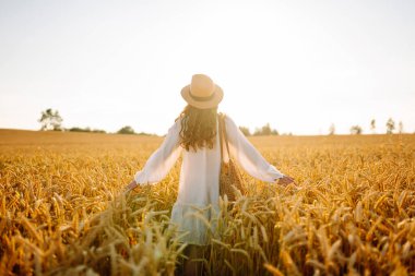 Şapkalı güzel bir kadın gün batımında altın buğday tarlasında yürüyor. Beyaz elbiseli mutlu kadın kırsal bir manzaranın tadını çıkarıyor. Güzellik, moda, hafta sonu konsepti. Yaşam biçimi.