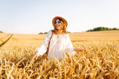 Şapkalı güzel bir kadın gün batımında altın buğday tarlasında yürüyor. Beyaz elbiseli mutlu kadın kırsal bir manzaranın tadını çıkarıyor. Güzellik, moda, hafta sonu konsepti. Yaşam biçimi.