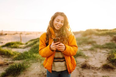 Sahile giden yolda telefonu olan kadın yürüyüşçünün portresi. Genç bir kadın gün doğumundan ve deniz manzaralı akıllı telefondan mesaj atmaktan hoşlanıyor. Macera ve teknoloji kavramı.