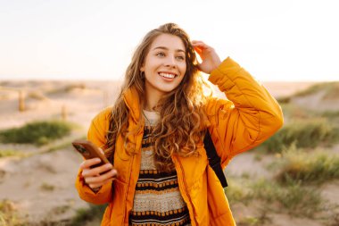 Sahile giden yolda telefonu olan kadın yürüyüşçünün portresi. Genç bir kadın gün doğumundan ve deniz manzaralı akıllı telefondan mesaj atmaktan hoşlanıyor. Macera ve teknoloji kavramı.