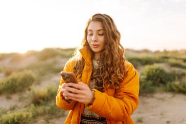 Sahile giden yolda telefonu olan kadın yürüyüşçünün portresi. Genç bir kadın gün doğumundan ve deniz manzaralı akıllı telefondan mesaj atmaktan hoşlanıyor. Macera ve teknoloji kavramı.