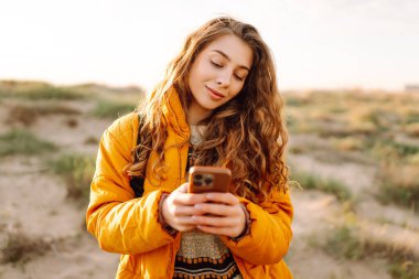Sahile giden yolda telefonu olan kadın yürüyüşçünün portresi. Genç bir kadın gün doğumundan ve deniz manzaralı akıllı telefondan mesaj atmaktan hoşlanıyor. Macera ve teknoloji kavramı.