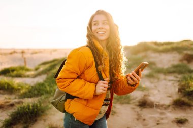 Sahile giden yolda telefonu olan kadın yürüyüşçünün portresi. Genç bir kadın gün doğumundan ve deniz manzaralı akıllı telefondan mesaj atmaktan hoşlanıyor. Macera ve teknoloji kavramı.
