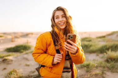 Sahile giden yolda telefonu olan kadın yürüyüşçünün portresi. Genç bir kadın gün doğumundan ve deniz manzaralı akıllı telefondan mesaj atmaktan hoşlanıyor. Macera ve teknoloji kavramı.