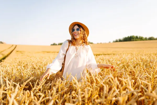Şapkalı güzel bir kadın gün batımında altın buğday tarlasında yürüyor. Beyaz elbiseli mutlu kadın kırsal bir manzaranın tadını çıkarıyor. Güzellik, moda, hafta sonu konsepti. Yaşam biçimi.