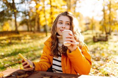 Parlak ceketli, telefonlu ve bir bardak kahveli iğrenç kadın sonbahar parkındaki ahşap bir masada oturuyor. Güzel bir kadın güneşli havadan hoşlanır ve sıcak bir içecek içer. Hafta sonu ve içki konsepti.