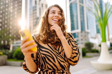 Telefonlu kıvırcık kadın, gün batımında metropolün arka planında poz veriyor. Şehir caddesinde yürürken akıllı telefon blogu yazan ya da mesajlaşan genç bir turist. Teknoloji ve blog kavramı.