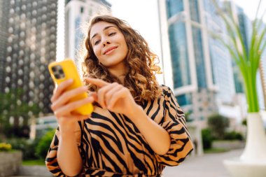 Telefonlu kıvırcık kadın, gün batımında metropolün arka planında poz veriyor. Şehir caddesinde yürürken akıllı telefon blogu yazan ya da mesajlaşan genç bir turist. Teknoloji ve blog kavramı.