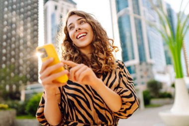 Telefonlu kıvırcık kadın, gün batımında metropolün arka planında poz veriyor. Şehir caddesinde yürürken akıllı telefon blogu yazan ya da mesajlaşan genç bir turist. Teknoloji ve blog kavramı.