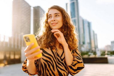 Telefonlu kıvırcık kadın, gün batımında metropolün arka planında poz veriyor. Şehir caddesinde yürürken akıllı telefon blogu yazan ya da mesajlaşan genç bir turist. Teknoloji ve blog kavramı.