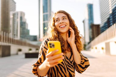 Telefonlu kıvırcık kadın, gün batımında metropolün arka planında poz veriyor. Şehir caddesinde yürürken akıllı telefon blogu yazan ya da mesajlaşan genç bir turist. Teknoloji ve blog kavramı.