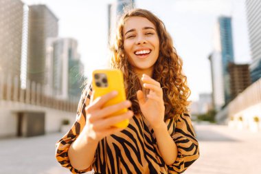 Telefonlu kıvırcık kadın, gün batımında metropolün arka planında poz veriyor. Şehir caddesinde yürürken akıllı telefon blogu yazan ya da mesajlaşan genç bir turist. Teknoloji ve blog kavramı.