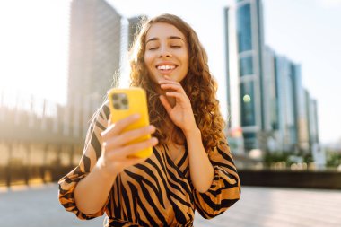 Telefonlu kıvırcık kadın, gün batımında metropolün arka planında poz veriyor. Şehir caddesinde yürürken akıllı telefon blogu yazan ya da mesajlaşan genç bir turist. Teknoloji ve blog kavramı.