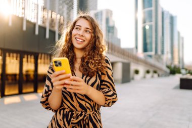 Telefonlu kıvırcık kadın, gün batımında metropolün arka planında poz veriyor. Şehir caddesinde yürürken akıllı telefon blogu yazan ya da mesajlaşan genç bir turist. Teknoloji ve blog kavramı.