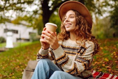 Şapkalı hoş bir kadın nehir kıyısında kahve fincanıyla kırmızı battaniyeye oturmuş dinleniyor. Güz havasının tadını çıkaran güzel bir kadın içkiyle parkta. Rahatlama, hafta sonu konsepti.