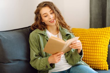 Gözlüklü ve kitaplı genç bir kadın rahat bir oturma odasında kanepede dinleniyor. Hoş bir kadın kitap okuyor ve huzurun tadını çıkarıyor. Öğrenme, okuma, rahatlama kavramı..