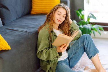 Gözlüklü ve kitaplı genç bir kadın rahat bir oturma odasında kanepede dinleniyor. Hoş bir kadın kitap okuyor ve huzurun tadını çıkarıyor. Öğrenme, okuma, rahatlama kavramı..