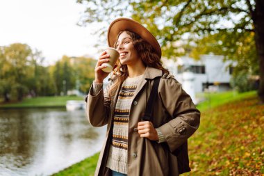 Bulutlu bir günde, şapkalı ve sıcak bir bardak içecekle parkta yürüyüş yapan genç bir kadın. Dışarıda kahve içen güzel bir kadın. Hafta sonu konsepti, turizm.