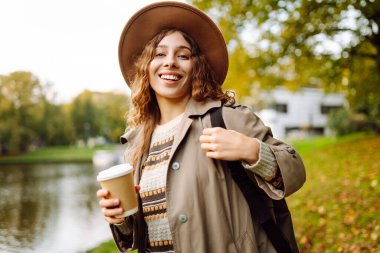 Bulutlu bir günde, şapkalı ve sıcak bir bardak içecekle parkta yürüyüş yapan genç bir kadın. Dışarıda kahve içen güzel bir kadın. Hafta sonu konsepti, turizm.