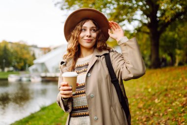 Bulutlu bir günde, şapkalı ve sıcak bir bardak içecekle parkta yürüyüş yapan genç bir kadın. Dışarıda kahve içen güzel bir kadın. Hafta sonu konsepti, turizm.