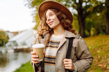 Bulutlu bir günde, şapkalı ve sıcak bir bardak içecekle parkta yürüyüş yapan genç bir kadın. Dışarıda kahve içen güzel bir kadın. Hafta sonu konsepti, turizm.