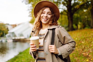 Bulutlu bir günde, şapkalı ve sıcak bir bardak içecekle parkta yürüyüş yapan genç bir kadın. Dışarıda kahve içen güzel bir kadın. Hafta sonu konsepti, turizm.