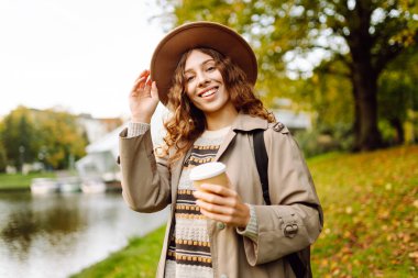 Bulutlu bir günde, şapkalı ve sıcak bir bardak içecekle parkta yürüyüş yapan genç bir kadın. Dışarıda kahve içen güzel bir kadın. Hafta sonu konsepti, turizm.