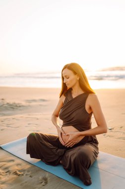 Yoga minderli hamile kadın kumlu sahilde rahatlama egzersizi yapıyor. Müstakbel anne güneş doğarken egzersiz yapıyor ve meditasyon yapıyor. Deniz manzarasının tadını çıkarıyor. Sağlık, rahatlama konsepti.