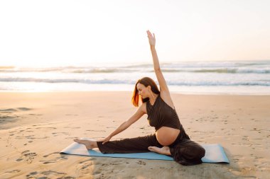 Yoga minderli hamile kadın kumlu sahilde rahatlama egzersizi yapıyor. Müstakbel anne güneş doğarken egzersiz yapıyor ve meditasyon yapıyor. Deniz manzarasının tadını çıkarıyor. Sağlık, rahatlama konsepti.