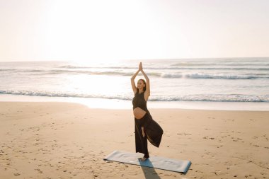 Yoga minderli hamile kadın kumlu sahilde rahatlama egzersizi yapıyor. Müstakbel anne güneş doğarken egzersiz yapıyor ve meditasyon yapıyor. Deniz manzarasının tadını çıkarıyor. Sağlık, rahatlama konsepti.