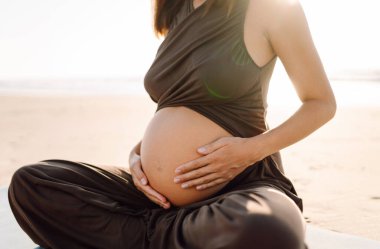 Yoga minderli hamile kadın kumlu sahilde rahatlama egzersizi yapıyor. Müstakbel anne güneş doğarken egzersiz yapıyor ve meditasyon yapıyor. Deniz manzarasının tadını çıkarıyor. Sağlık, rahatlama konsepti.