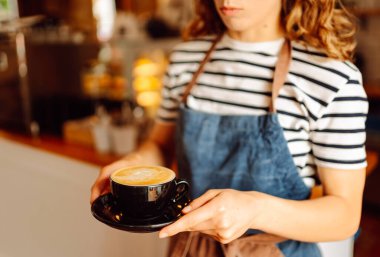 Kahve dükkanının tezgahında elinde bir fincan sıcak kahve tutan bayan baristanın yakın çekimi. Genç kadın Inapron rahat bir yerde güzel kokulu bir kahve içeceği hazırladı. Küçük işletme, içecek konsepti.