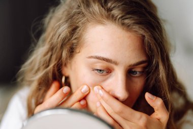 Kıvırcık kadın cildini inceler ve küçük aynaya bakarken elleriyle dokunur. Doğal güzellik. Şirin bir kadın sıcak bir odada kızarıklığı parmaklarıyla işaret ediyor. Güzellik ve kozmetoloji kavramı.