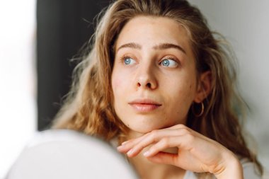 Kıvırcık kadın cildini inceler ve küçük aynaya bakarken elleriyle dokunur. Doğal güzellik. Şirin bir kadın sıcak bir odada kızarıklığı parmaklarıyla işaret ediyor. Güzellik ve kozmetoloji kavramı.