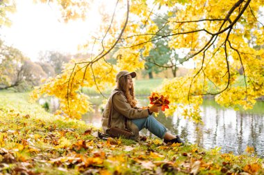 Mutlu kadın güneşli bir sonbahar parkında bir buket parlak yaprakla eğleniyor. Günbatımının tadını çıkaran günlük giysiler içinde güzel bir turist. Yürüme konsepti. Sonbahar atmosferi.