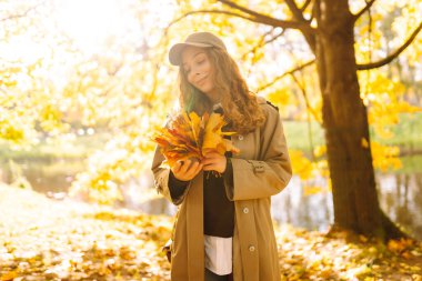 Mutlu kadın güneşli bir sonbahar parkında bir buket parlak yaprakla eğleniyor. Günbatımının tadını çıkaran günlük giysiler içinde güzel bir turist. Yürüme konsepti. Sonbahar atmosferi.