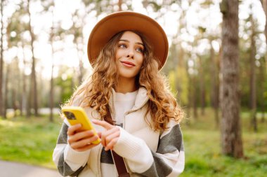 Telefonlu ve şapkalı genç bir kadın güneşli bir parkta eğleniyor. Güzel bir kadın gün batımında yürümekten ve mesajlaşmaktan zevk alıyor. Blog yazma, boş zaman konsepti. Etkin yaşam biçimi.