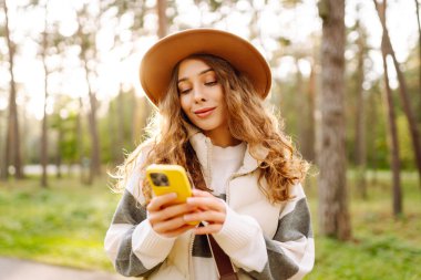 Telefonlu ve şapkalı genç bir kadın güneşli bir parkta eğleniyor. Güzel bir kadın gün batımında yürümekten ve mesajlaşmaktan zevk alıyor. Blog yazma, boş zaman konsepti. Etkin yaşam biçimi.