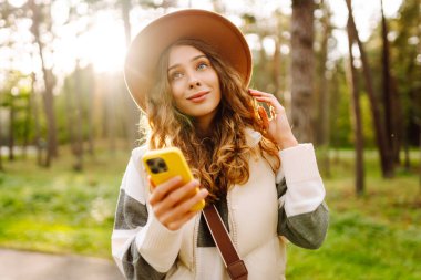 Telefonlu ve şapkalı genç bir kadın güneşli bir parkta eğleniyor. Güzel bir kadın gün batımında yürümekten ve mesajlaşmaktan zevk alıyor. Blog yazma, boş zaman konsepti. Etkin yaşam biçimi.