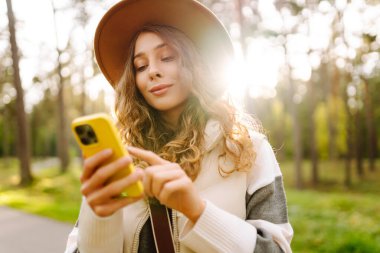 Telefonlu ve şapkalı genç bir kadın güneşli bir parkta eğleniyor. Güzel bir kadın gün batımında yürümekten ve mesajlaşmaktan zevk alıyor. Blog yazma, boş zaman konsepti. Etkin yaşam biçimi.